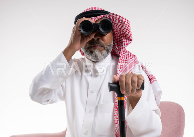 الجلوس على الكرسي بإيماءات التركيز والاستكشاف, بورتريه مقرب لرجل كبير بالسن عربي خليجي سعودي يرتدي الثوب والتقليدي والشماغ يمسك بيده العكاز ويقوم بالنظر عبر منظار التلسكوب ثنائي العينين, رؤية الأشياء البعيدة, خلفية بيضاء