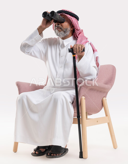 حب الاستكشاف والمغامرة, تحقيق الأهداف والطموحات المستقبلية, استخدام ناظور ثنائي العينين التلسكوب لرؤية الأشياء البعيدة, بورتريه لرجل كبير بالسن عربي خليجي سعودي يرتدي الثوب التقليدي والشماغ يمسك بيده العكاز وينظر إلى شيء ما عبر منظار الرؤية, خلفية بيضاء