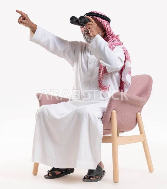 الجلوس والتخطيط المستقبلي للأهداف والطموحات, بورتريه لرجل شايب عربي خليجي سعودي يرتدي الثوب التقليدي والشماغ ينظر عبر منظار الرؤية ويشير بيده إلى شيء ما, رؤية الأشياء البعيدة عبر ناظور التلسكوب, حب الاستكشاف والمغامرة, خلفية بيضاء