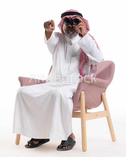 التطلع والنظرة المستقبلية, مفهوم المغامرة والاستكشاف, بورتريه لرجل شايب عربي خليجي سعودي يرتدي الثوب التقليدي والشماغ ينظر من خلال منظار الرؤية ويشير بإصبعه إلى الكاميرا, رؤية الأشياء البعيدة عبر ناظور ثنائي العينين التلسكوب, خلفية بيضاء