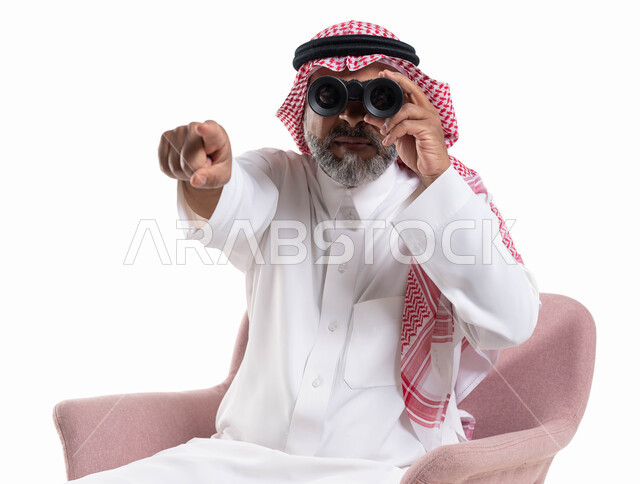 مفهوم المغامرة والاستكشاف, بورتريه مقرب لرجل شايب عربي خليجي سعودي يرتدي الثوب التقليدي والشماغ ينظر من خلال منظار الرؤية ويشير بإصبعه إلى الكاميرا, رؤية الأشياء البعيدة عبر ناظور ثنائي العينين التلسكوب, التطلع والنظرة المستقبلية, خلفية بيضاء