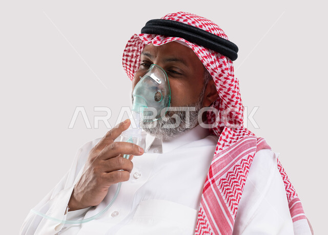 معالجة أمراض الربو المزمنة، المحافظة على ممر هوائي منتظم باستخدام جهاز التنفس الصناعي، بورتريه مقرب لرجل كبير بالسن عربي سعودي خليجي يرتدي الشماغ والثوب التقليدي يقوم باستنشاق أدوية الكحة، تقديم الخدمات الصحية المنزلية للمريض، خلفية بيضاء