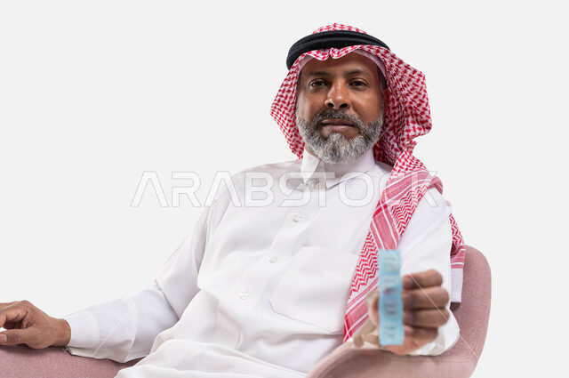 الاهتمام بالصحة والالتزام بالأوقات الصحيحة لأخذ العلاج, استخدام عقار الدواء بوصفة طبية, بورتريه مقرب لرجل كبير بالسن عربي خليجي سعودي يرتدي الثوب التقليدي والشماغ يجلس على الكرسي يحمل شريط الدواء, مفهوم الطب والرعاية الطبية, خلفية بيضاء