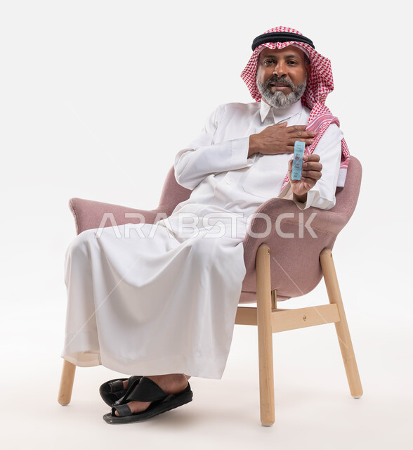 استخدام عقار الدواء بوصفة طبية, بورتريه لرجل كبير بالسن عربي خليجي سعودي يرتدي الثوب التقليدي والشماغ يجلس على الكرسي يحمل شريط الدواء, مفهوم الطب والرعاية الطبية, الاهتمام بالصحة والالتزام بالأوقات الصحيحة لأخذ العلاج, خلفية بيضاء