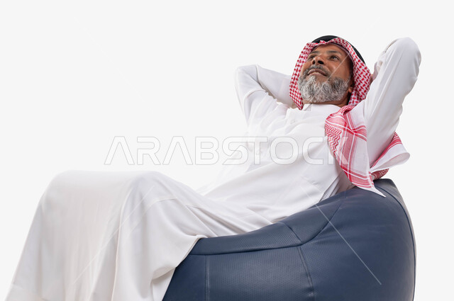 الجلوس براحة والنظر إلى شيء ما بإيماءات التأمل والتفكير وشرود الذهن، بورتريه لرجل عربي خليجي سعودي كبير بالسن يرتدي الشماغ والثوب التقليدي يضع يديه تحت رأسه ويستلقي على كرسي بنظرة وتطلع للمستقبل، خلفية بيضاء