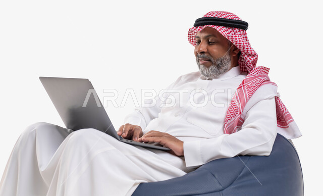انجاز الاعمال والمهام عن بعد عبر الحاسوب المحمول، تصفح الانترنت ووسائل التواصل الاجتماعي، بورتريه مقرب لرجل عربي سعودي خليجي كبير بالسن يرتدي الشماغ والثوب التقليدي يجلس على كرسي مريح يقوم باستخدام الحاسب، خلفية بيضاء