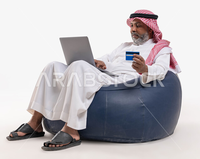 Providing means to help the elderly in the purchase and payment process remotely, using a credit card in electronic shopping, a portrait of a Saudi Gulf Arab man wearing a shemagh and traditional dress, sitting in a comfortable chair holding a bank card and using a laptop, white background.