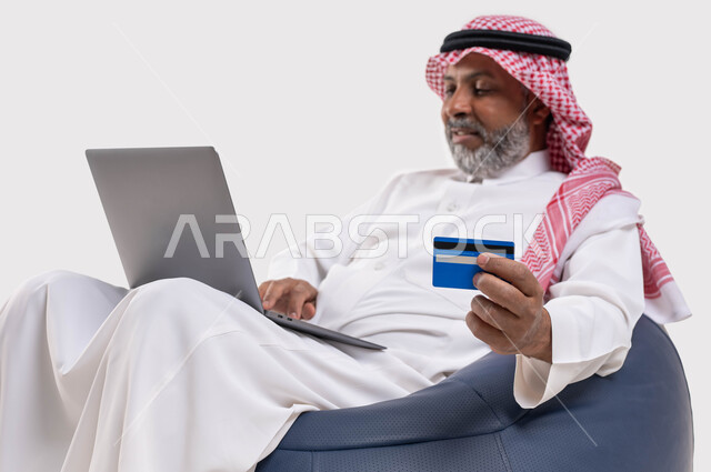 إدخال المعلومات البنكية على الحاسوب المحمول، توفير وسائل تساعد كبار السن في عملية الشراء والدفع عن بعد، بورتريه مقرب لرجل عربي سعودي خليجي يرتدي الشماغ والثوب التقليدي يجلس على كرسي مريح يقوم باستخدام بطاقة الإئتمان في التسوق الالكتروني عبر الحاسب، خلفية بيضاء