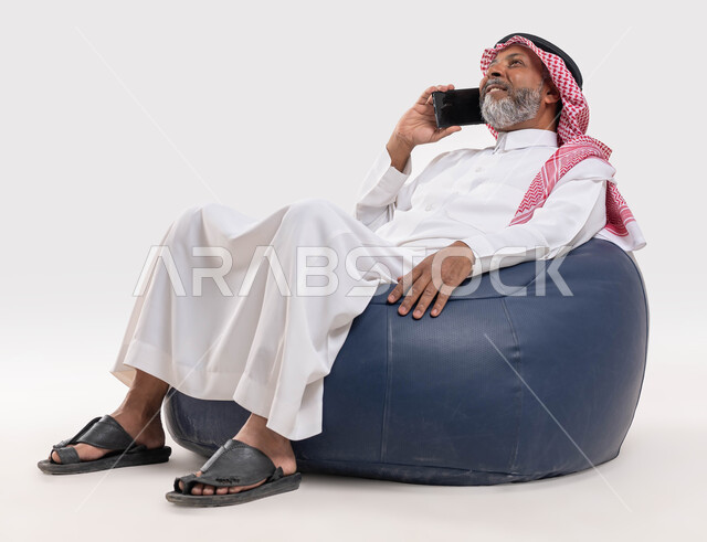 الرد على الاتصالات والمكالمات، سماع أخبار سعيدة عبر الهاتف المحمول، بورتريه لرجل عربي سعودي خليجي كبير بالسن يرتدي الشماغ والثوب التقليدي يجلس على كرسي مريح يقوم بإجراء مكالمة هاتفية، التواصل مع الأصدقاء والاقارب باستخدام الجوال، خلفية بيضاء