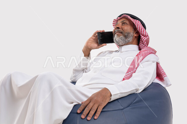 التواصل مع الأصدقاء والاقارب باستخدام الجوال، الرد على الاتصالات والمكالمات، سماع أخبار سعيدة عبر الهاتف المحمول، بورتريه مقرب لرجل عربي سعودي خليجي كبير بالسن يرتدي الشماغ والثوب التقليدي يجلس على كرسي مريح يقوم بإجراء مكالمة هاتفية، خلفية بيضاء