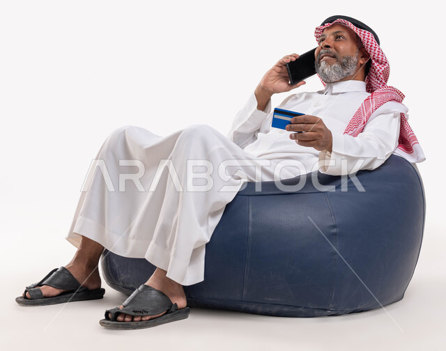 Providing means to help the elderly in the process of purchasing and paying remotely, using a credit card in electronic shopping, a portrait of a Saudi Arabian Gulf man wearing a shemagh and a traditional thobe, sitting in a comfortable chair holding a bank card, making a phone call via a mobile phone, white background.