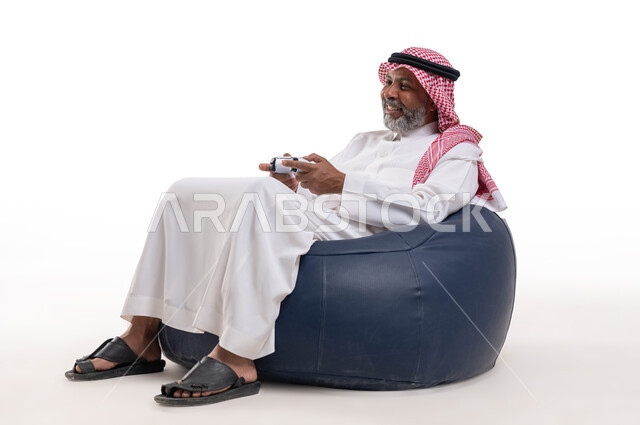 تقنيات الكترونية حديثة ترفيهيه, قضاء وقت ممتع ومسلي، التفاعل والاندماج مع ألعاب البلايستيشن، بورتريه لرجل عربي سعودي خليجي كبير بالسن يرتدي الشماغ والثوب التقليدي يجلس على كرسي مريح يحمل بيده عصا التحكم يقوم باللعب في ألعاب الفيديو، خلفية بيضاء
