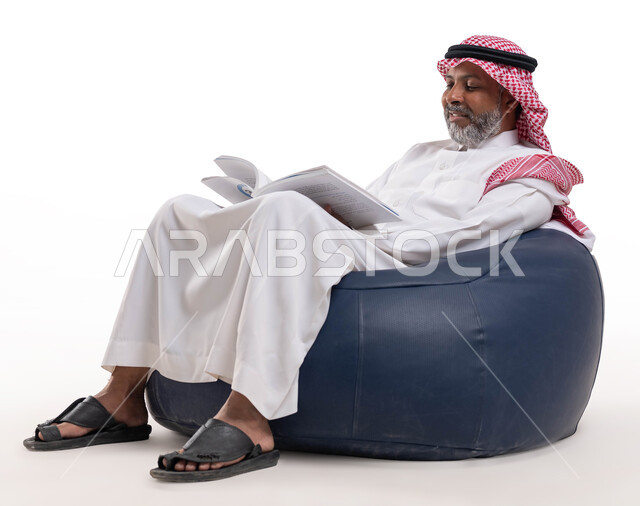 القراءة بتمعن وتركيز، قضاء وقت الفراغ بالتعلم والثقافة، مفهوم محو الامية، بورتريه لرجل كبير بالسن عربي سعودي خليجي مبتسم يرتدي الشماغ والثوب التقليدي يجلس على كرسي مريح يحمل بيده كتاب علمي تثقيفي، خلفية بيضاء