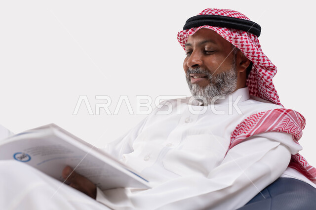 Reading with concentration and thought, spending your free time with learning and culture, the concept of eradicating illiteracy, a close-up portrait of an elderly, smiling Saudi Arabian Gulf man wearing a shemagh and traditional thobe, sitting on a comfortable chair holding an educational scientific book in his hand, white background.