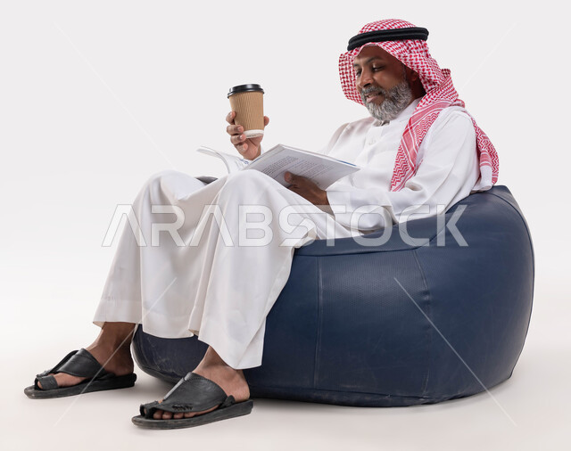 قضاء وقت الفراغ بالتعلم والثقافة، مفهوم محو الامية، القراءة بتمعن وتركيز، بورتريه لرجل كبير بالسن عربي سعودي خليجي مبتسم يرتدي الشماغ والثوب التقليدي يجلس على كرسي مريح يحمل بيده كوب مشروب ساخن وكتاب تثقيفي، خلفية بيضاء
