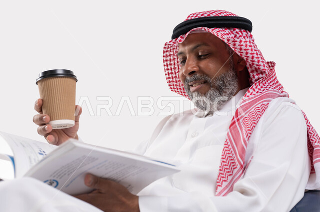 مفهوم محو الامية، قضاء وقت الفراغ بالتعلم والثقافة، القراءة بتمعن وتركيز، بورتريه مقرب لرجل عربي سعودي خليجي كبير بالسن مبتسم يرتدي الشماغ والثوب التقليدي يجلس على كرسي مريح يحمل بيده كوب مشروب ساخن وكتاب تثقيفي، خلفية بيضاء
