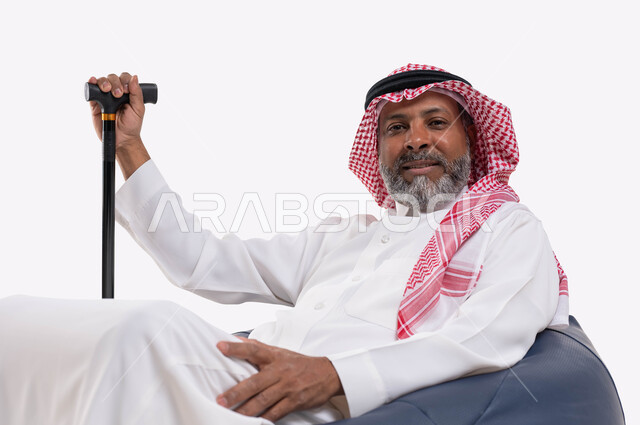 The concept of sitting and resting, a close-up portrait of an elderly Saudi Gulf Arab man, smiling, wearing a shemagh and traditional dress, sitting on a comfortable chair holding a crutch in his hand, looking at the camera with self-confidence gestures, white background.