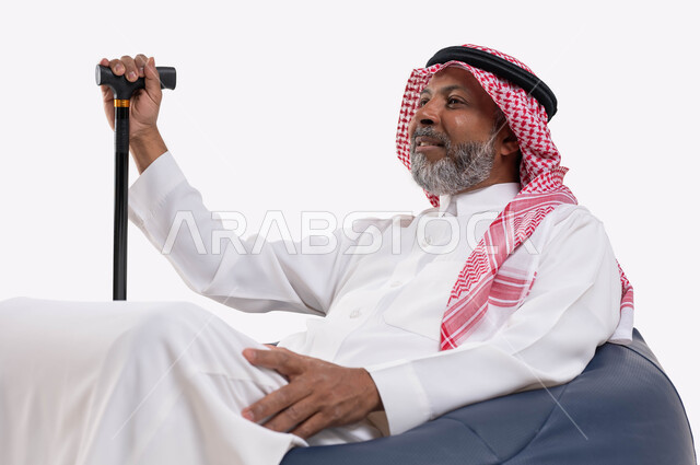 إيماءات التفكير والشرود، مفهوم الجلوس والراحة، بورتريه مقرب لرجل عربي خليجي سعودي مبتسم كبير بالسن يرتدي الشماغ والثوب التقليدي يجلس على كرسي مريح يمسك بيده العكاز، النظر الى شيء ما، خلفية بيضاء
