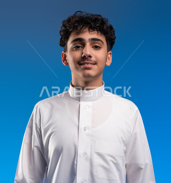 The concept of masculinity and elegance, attention to external appearance, a close-up portrait of a smiling Saudi Gulf Arab young man wearing a traditional dress, standing straight and looking at the camera with happy gestures, blue background.