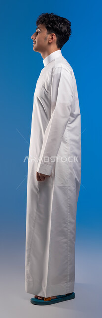 The concept of elegance and attention to external appearance, standing confidently and straight, portrait from the side of a smiling Saudi Gulf Arab man wearing traditional clothing looking at something, full-length body portrait, blue background