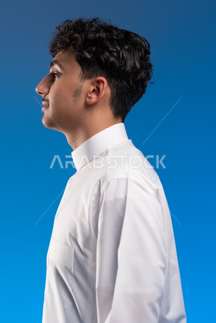 الوقوف بثقة واستقامة، بورتريه مقرب من الجانب لشاب عربي خليجي سعودي مبتسم يرتدي الزي التقليدي ينظر إلى شيء ما، مفهوم الاناقة والاهتمام بالمظهر الخارجي، خلفية زرقاء
