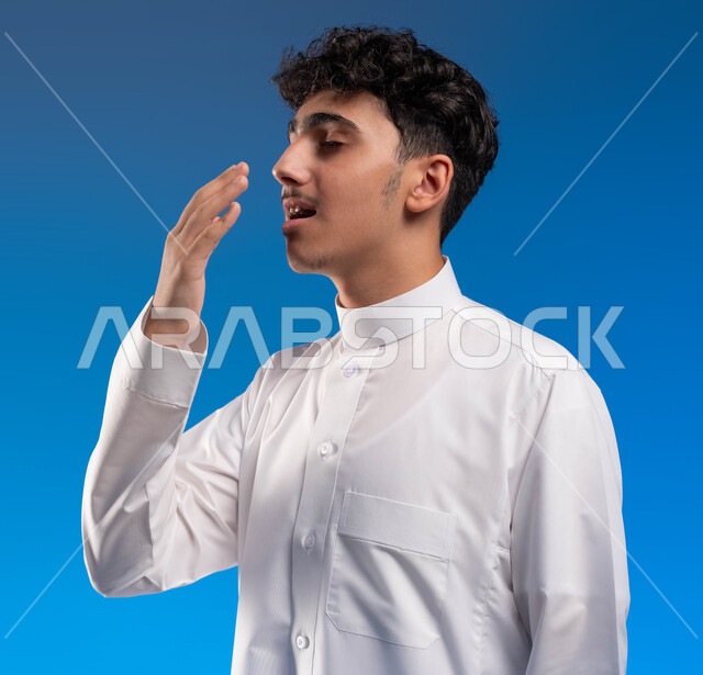 الشعور بالتعب والتثاؤب و الكسل والخمول، الإحساس بالإرهاق و الحاجه الى النوم والراحه ,بورتريه مقرب لشاب عربي خليجي سعودي يرتدي الزي التقليدي يضع يده على فمه ،إيماءات تدل على النعاس, خلفية زرقاء
