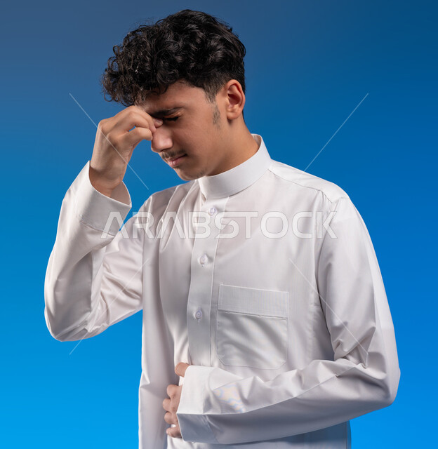 Feelings of headache and illness, feeling of dizziness and imbalance, close-up portrait of a young Saudi Arabian Gulf man wearing traditional clothing, placing his hand on his face, fatigue and exhaustion, and the need for treatment, asking for help to go to the hospital, blue background.