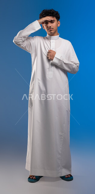 إيماءات وجه تدل على الشعور بالصداع، الاحساس بالمرض و ألم بالرأس, بورتريه لشاب عربي سعودي خليجي يرتدي الزي التقليدي ينظر إلى الكاميرا، التعب والإرهاق و الحاجه للعلاج, طلب المساعده للذهاب الى المستشفى, صورة للجسم بالكامل، خلفية زرقاء