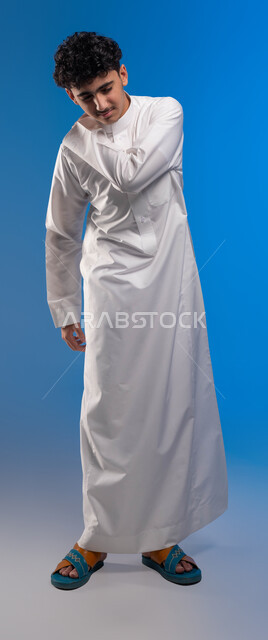 طلب المساعده للذهاب الى المستشفى، النظر إلى الكاميرا، شد عضلي في الرقبه ،التهاب العضلات والمفاصل، الإحساس بالتعب والإرهاق, بورتريه مقرب لشاب عربي سعودي خليجي يضع يده على رقبته بإيماءات الألم في الأكتاف، صورة الجسم بالطول الكامل، خلفية زرقاء.