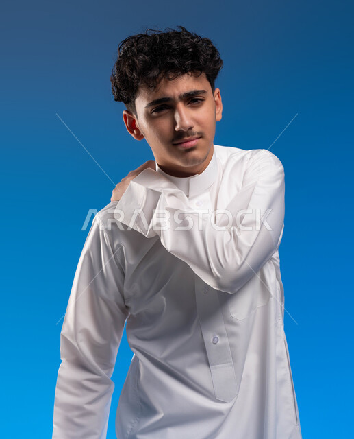 النظر إلى الكاميرا، شد عضلي في الرقبه ،التهاب العضلات والمفاصل، الإحساس بالتعب والإرهاق, بورتريه مقرب لشاب عربي سعودي خليجي يضع يده على رقبته بإيماءات الألم في الأكتاف، الحاجه للعلاج و وضع خطة علاجية, طلب المساعده للذهاب الى المستشفى، خلفية زرقاء.
