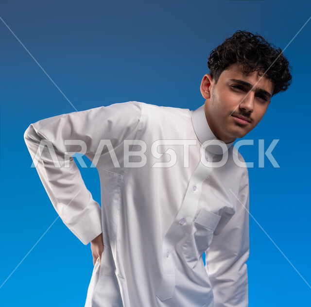 طلب المساعده للذهاب الى الطبيب , التعب والإرهاق و الحاجه للعلاج, الإحساس بالمرض ووجع العظام والعضلات, بورتريه مقرب لشاب عربي خليجي سعودي يرتدي الزي التقليدي وينحني شعورا بالألم في الظهر, خلفية زرقاء
