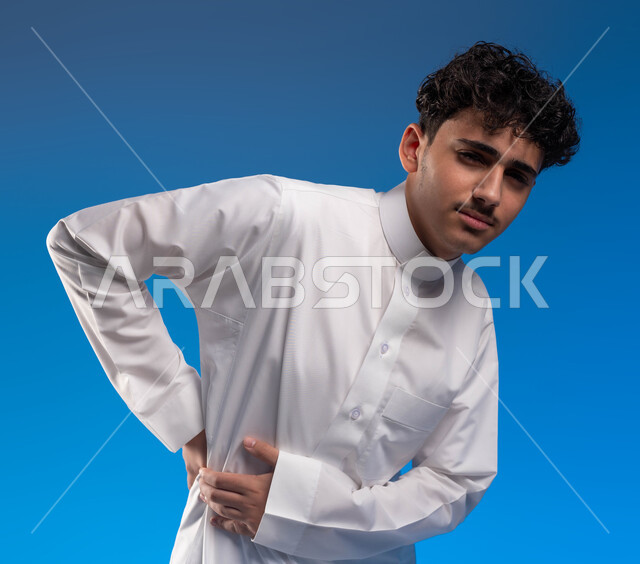 النظر إلى الكاميرا، الاحساس بألم في العمود الفقري , بورتريه مقرب لشاب عربي خليجي سعودي يرتدي الزي التقليدي وينحني شعورا بأوجاع في أسفل الظهر, التعب والإرهاق و الحاجه للعلاج, طلب المساعده للذهاب الى الطبيب , خلفية زرقاء