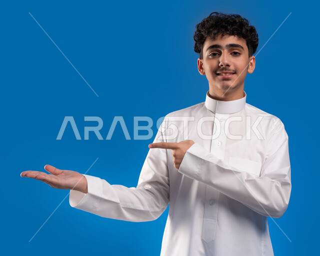 تحديد شيء معين بإيماءات السرور, النظر إلى الكاميرا، بورتريه مقرب لشاب عربي خليجي سعودي مبتسم يرتدي الثوب التقليدي يفتح كفه للأعلى ويشير إلى عنصر ما, خلفيه زرقاء