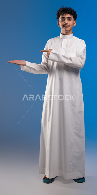 النظر إلى الكاميرا وتحديد شيء معين بإيماءات السرور, بورتريه لشاب عربي خليجي سعودي مبتسم يرتدي الثوب التقليدي يفتح كفه للأعلى ويشير إلى عنصر ما, صورة الجسم بالطول الكامل, خلفية زرقاء
