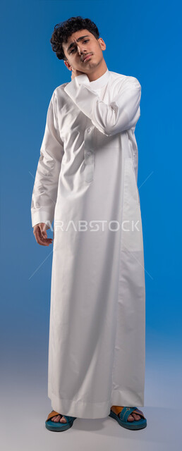 Gestures of pain in the shoulders, inflammation of the muscles and joints, a feeling of fatigue and exhaustion, a close-up portrait of a young Saudi Arabian Gulf man putting his hand on his neck and looking at the camera, the need for treatment and developing a treatment plan, asking for help to go to the hospital, full body photo, blue background