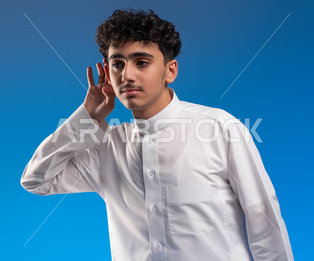 سماع الأسرار والمحادثات، محاولة التجسس والتنصت, بورتريه مقرب لشاب عربي سعودي خليجي يرتدي الزي التقليدي ويضع يده خلف اذنه, إيماءات تدل على الإنصات, حسن الإستماع و التركيز على الكلام, خلفية زرقاء