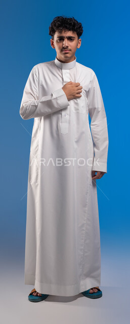 الشعور في زيادة معدل ضربات القلب، ضرورة الذهاب إلى الطبيب لإجراء الفحوصات اللازمة، اضطرابات و أمراض قلبية، بورتريه لشاب عربي خليجي سعودي يرتدي الزي التقليدي  يضع يده على صدره يشعر بحالة من الوخز والألم بقلبه , صورة الجسم بالطول الكامل، خلفية زرقاء