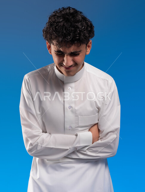 Facial movements indicating a feeling of pain, a feeling of cramping, fatigue and exhaustion, asking for help to go to the doctor, indigestion and a feeling of heartburn, a close-up portrait of a young Saudi Arabian Gulf man wearing traditional clothing holding his stomach with crossed hand gestures, blue background.