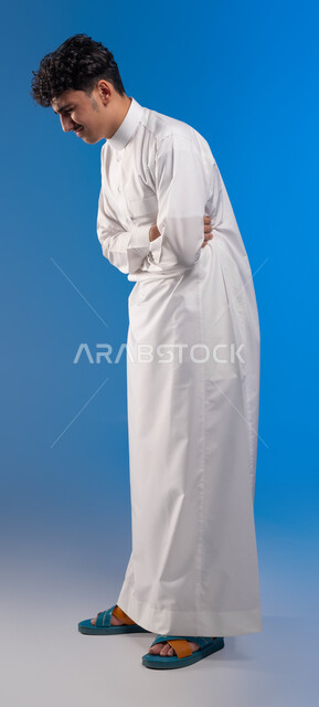 Feeling of cramps, fatigue and exhaustion, asking for help to go to the doctor, indigestion and feeling of heartburn, close-up portrait of a young Saudi Arabian Gulf man wearing traditional clothing holding his stomach with crossed hand gestures feeling pain, the need to develop a treatment plan, full body image, blue background