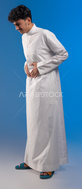 طلب المساعده للذهاب الى الطبيب، عسر الهضم والشعور بحرقة في المعدة، بورتريه لشاب عربي سعودي خليجي يرتدي الزي التقليدي يمسك بطنه شعورا بالألم، الإحساس بالمغص و التعب والإرهاق، الحاجة للدواء ووضع خطة علاجية، صورة الجسم بالكامل، خلفية زرقاء