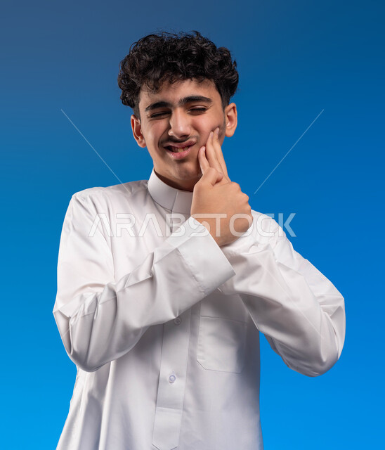 الحاجه لزيارة طبيب الأسنان لوضع خطة علاجية، بورتريه مقرب لشاب عربي خليجي سعودي يرتدي الزي التقليدي يضع يده على وجهه بإيماءات الألم، تسوس الأضراس و إلتهاب اللثه, تعابير الارهاق والتعب، خلفية زرقاء