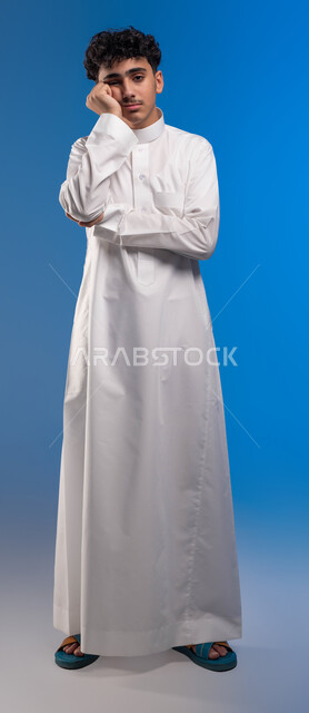 إيماءات التفكير والتأمل، الوقوف باستقامة والنظر إلى الكاميرا، بورتريه لشاب عربي سعودي خليجي يرتدي الثوب التقليدي يضع يده على وجهه، الشعور بالملل والكآبة، الحيرة والانتظار لأمر معين، صورة الجسم بالطول الكامل، خلفية زرقاء.