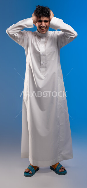 الشعور بالصداع و ألم بالرأس, الوقوف باستقامة والنظر إلى الكاميرا، الإحساس بالدوار والتعب والإرهاق, بورتريه مقرب لرجل عربي سعودي خليجي يرتدي الثوب التقليدي يضع يديه على رأٍسه بإيماءات الوجع, الحاجه للعلاج و طلب المساعده, صورة الجسم بالطول الكامل، خلفية زرقاء .