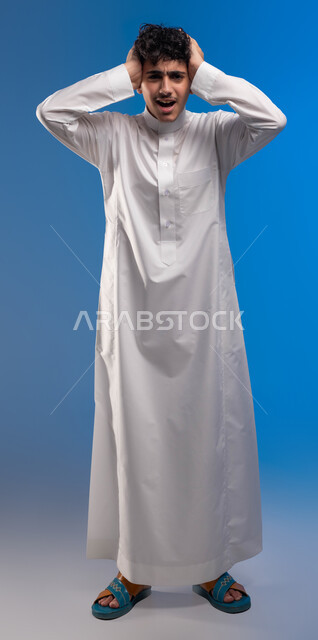 الوقوف باستقامة والنظر إلى الكاميرا، الإحساس بالدوار والتعب والإرهاق, بورتريه مقرب لرجل عربي سعودي خليجي يرتدي الثوب التقليدي يضع يديه على رأٍسه بإيماءات الوجع, الشعور بالصداع و ألم بالرأس, الحاجه للعلاج و طلب المساعده, صورة الجسم بالطول الكامل، خلفية زرقاء .
