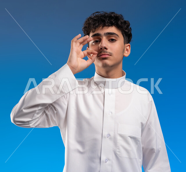 ايماءات الرجوله والثقة والقوة , الاعجاب بنمو الشارب، التهديد والتوعد لشيء ما، بورتريه مقرب لشاب عربي خليجي سعودي يرتدي الزي التقليدي ينظر إلى الكاميرا يمسك شاربه بيده, حركات وتعابير شبابيه،  صورة الجسم بالطول الكامل، خلفية زرقاء .