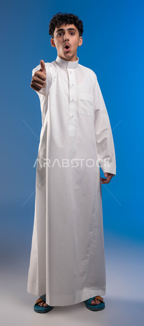 إيماءات الذهول والاندهاش، الشعور بالموافقه والقبول والرضا, بورتريه مقرب لشاب عربي خليجي سعودي يرتدي الثوب التقليدي يرفع أصبع الإبهام نحو الأعلى بعلامة الجودة، تعابير الحماس والفرح، الإمتياز والتميز و النجاح, صورة الجسم بالطول الكامل، خلفية زرقاء .