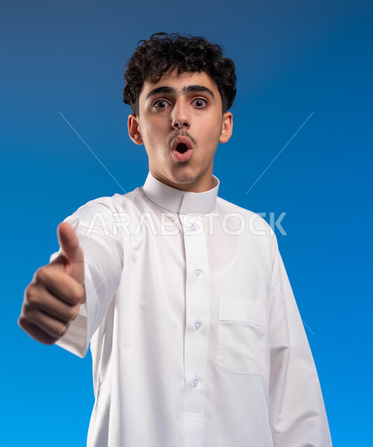 الشعور بالموافقه والقبول والرضا, بورتريه مقرب لشاب عربي خليجي سعودي يرتدي الثوب التقليدي يرفع أصبع الإبهام نحو الأعلى بعلامة الجودة، إيماءات الذهول والاندهاش، تعابير الحماس والفرح، الإمتياز والتميز و النجاح, خلفية زرقاء .
