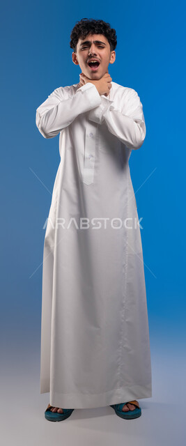 الاصابة بنزلة برد والتهاب الحلق، إيماءات صعوبة التنفس والتعب والألم، بورتريه شاب عربي سعودي خليجي يرتدي الثوب التقليدي يضع يديه على حلقه يشعر بالاختناق، المرض الشديد والسعال، العدوى بفايروس كورونا، صورة الجسم الطول الكامل، خلفية زرقاء .
