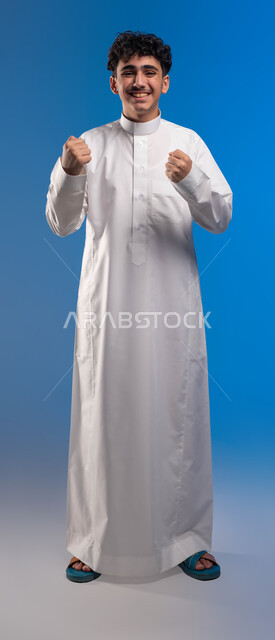 الشعور بالقوة والنجاح، إيماءات التفاعل والإندماج, بورتريه لشاب عربي سعودي خليجي يرتدي الزي السعودي يرفع يديه مضموم الكفين يقوم بالتشجيع, إيماءات السعادة والفوز والإنتصار, أنديه وتشجيع, صورة الجسم بالطول الكامل، خلفية زرقاء .