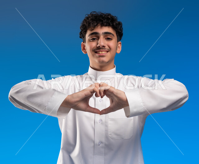 الاحتفال بالأعياد والمناسبات، بورتريه مقرب لشاب عربي سعودي خليجي مبتسم يرتدي الثوب التقليدي يشكل بيده شكل القلب، الشعور بالحب والفرح والسعادة، مشاعرالحب والغرام, إيماءات الاهتمام والعواطف، خلفية زرقاء .
